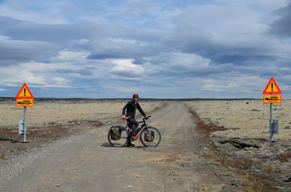 Fietser op een Santos reisfiets op een onverharde hooglandweg in IJsland, tussen twee verkeersborden die het traject als alleen begaanbaar voor 4x4-voertuigen markeren